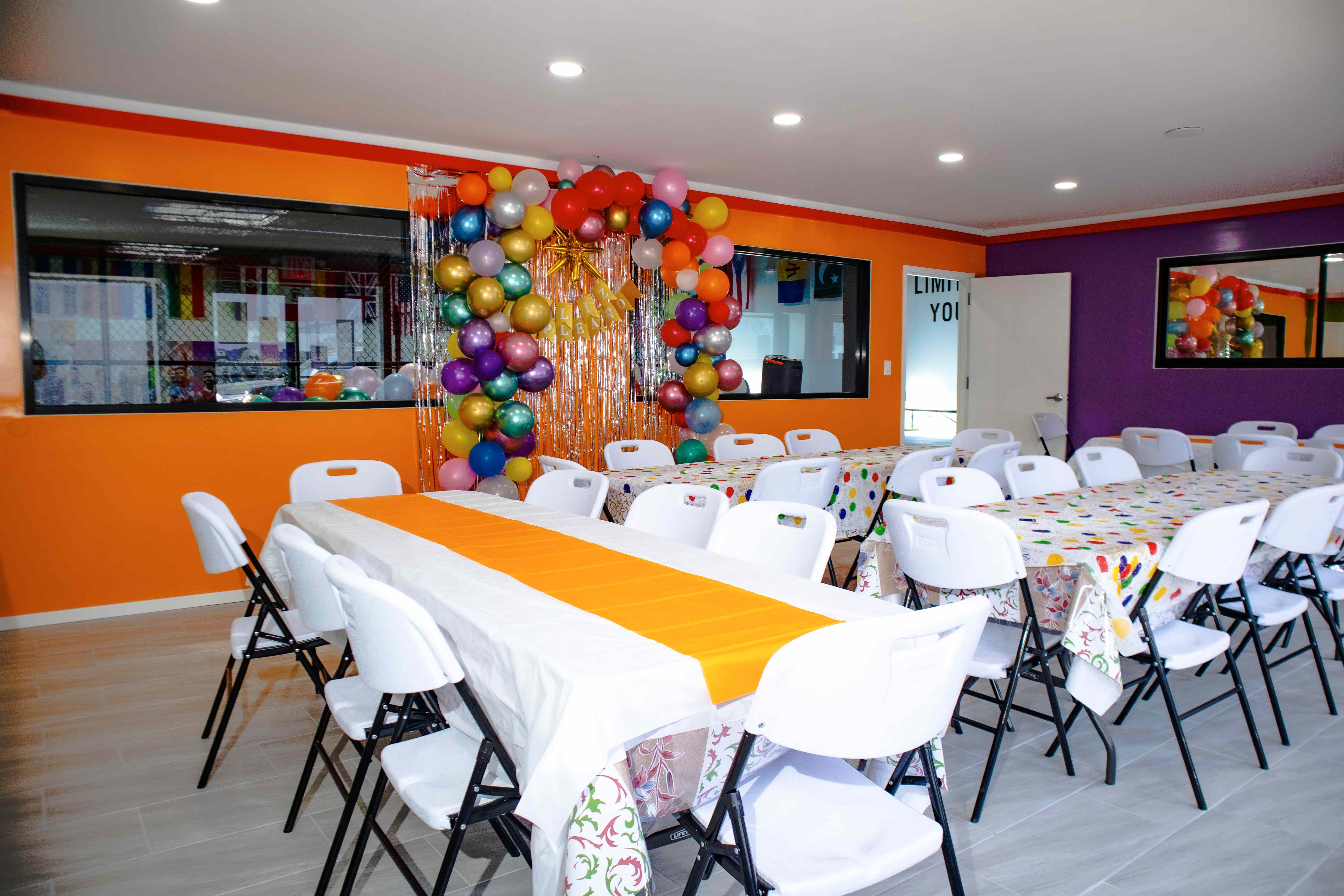 Party room with orange and purple walls, balloon arch, and tables set with white cloths