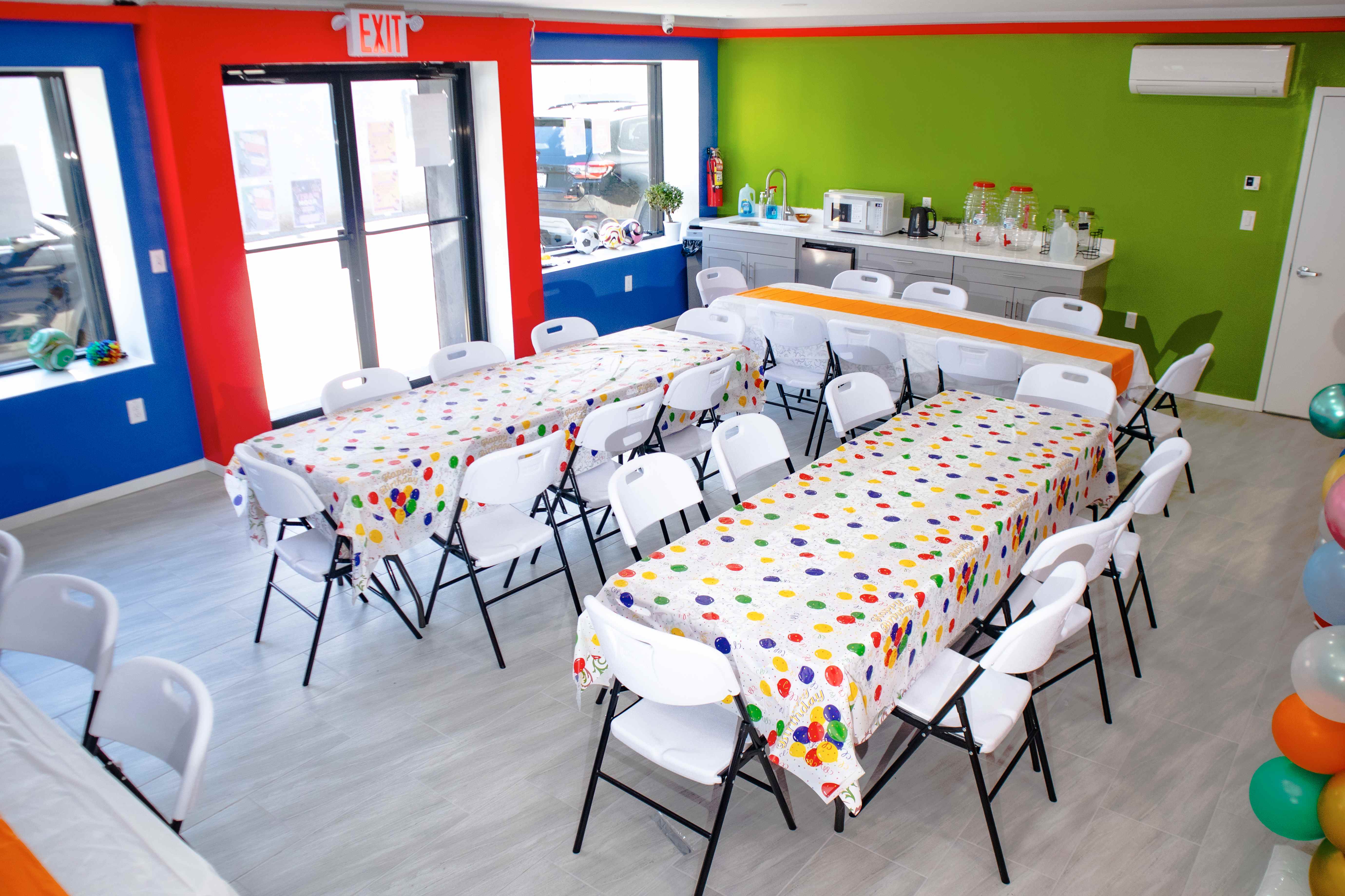 Bright party room with red, blue, and green walls, balloon-print tables, and a kitchenette