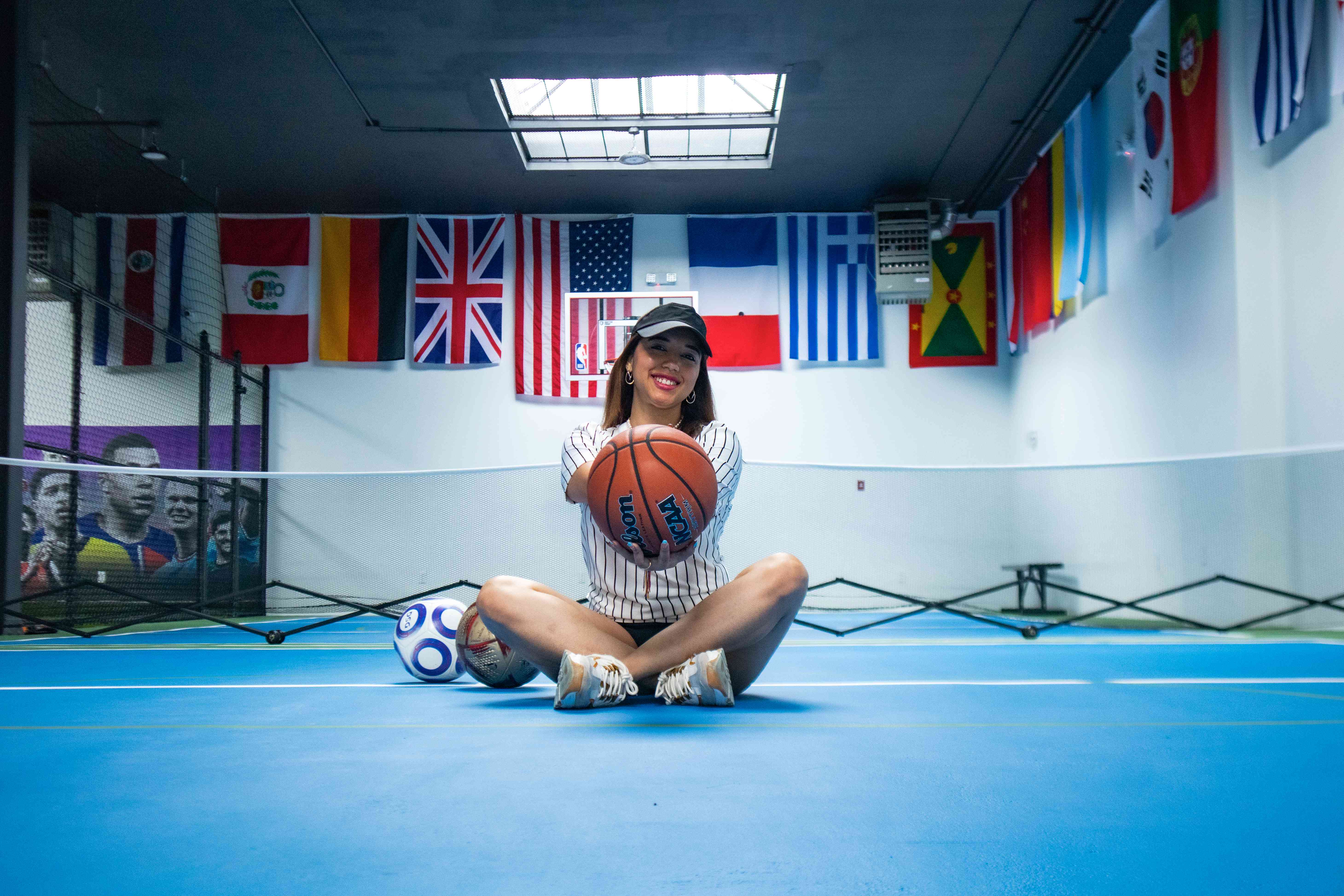 Player sitting on the court holding a basketball with soccer balls beside her